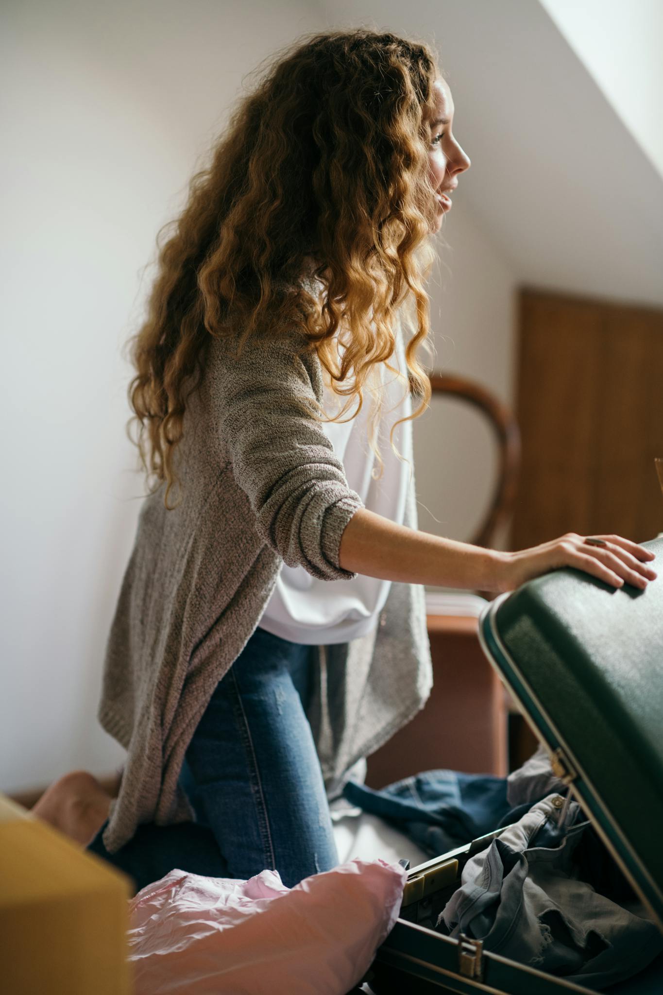 A young woman with long hair packs a suitcase in a cozy bedroom setting.
